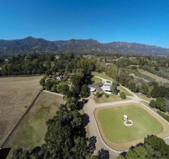 This drone aerial view shows the equestrian estate, including riding facilities, barn, and main residence, spread before the Santa Ynez Mountains.