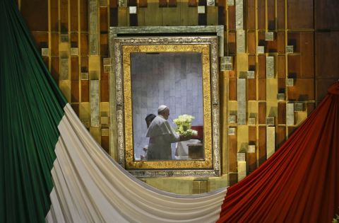 Pope Francis, reflected in the frame that holds the image of the Virgin of Guadalupe, holds a bouquet of flowers as he prays to her inside the basilica built in her honor.