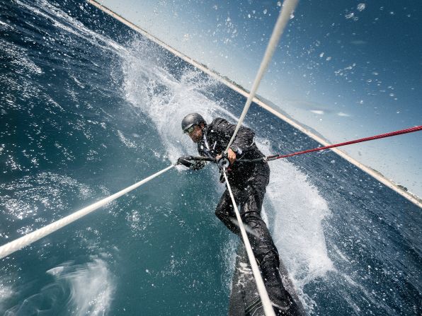 Alex Thomson begins his stunt by chasing his speeding IMOCA Open 60 yacht on a kiteboard, while dressed in a sharp suit and tie.