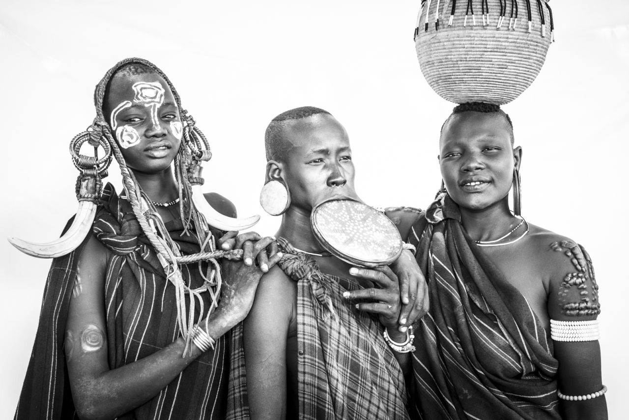 The scarification on shoulder of the woman standing on the right is prominent. Denoting beauty, the process involves cutting the skin with a razor and applying ash from fires to infect the wound. The scar tissue then causes the skin to protrude.