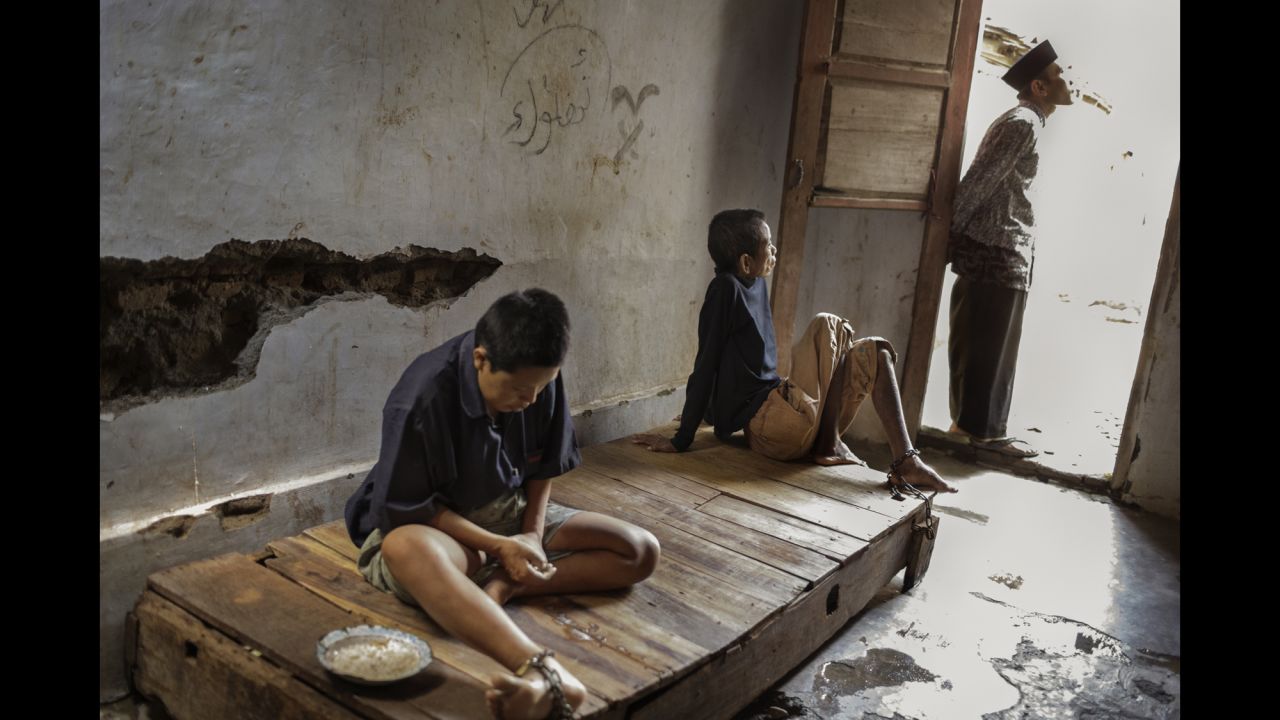 Two residents at the Bina Lestari healing center in Brebes, Central Java, are chained to a wooden platform bed while an Islamic faith healer stands nearby. At the center, all residents are chained and receive traditional "healing" through prayer, consumption of special drinks, or blessings with holy water.