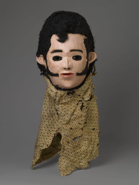 "Elvis Mask for Nyau Society", Unidentified Chewa artist (c. 1977). This wooden mask is from the Nyau Society, Malawi, whose masks represent spirits of the dead, wild bush spirits and caricatures of outsiders like Swahili slave traders, the Virgin Mary, and  iconic foreigners such as Elvis Presley.