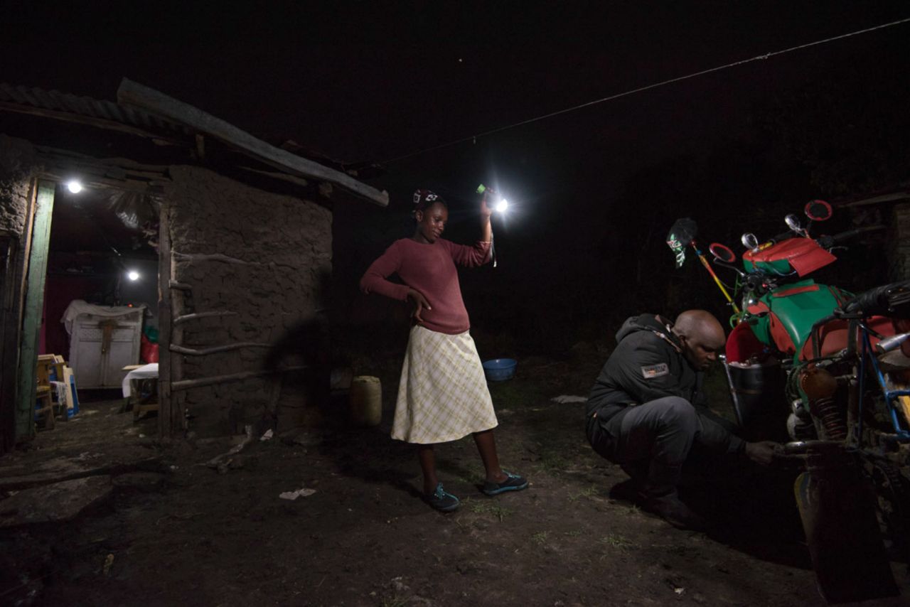 A woman lights up the compound as her husband fixes a loose screw on his motorbike. The lamp is powered by a solar panel which they have bought on M-KOPA's payment plan. 