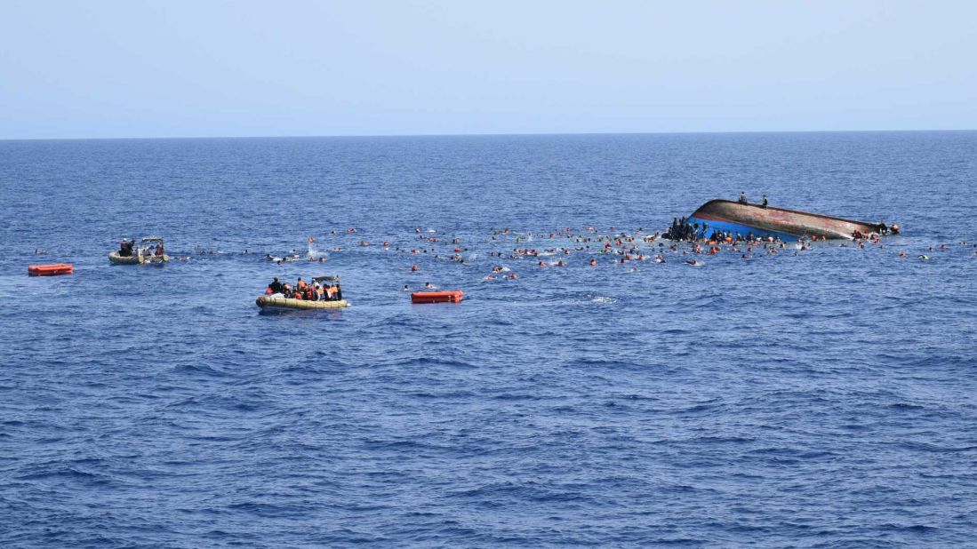 Migrants cling to the hull of the ship and float in the water as Italian sailors rescue them with life rafts. 