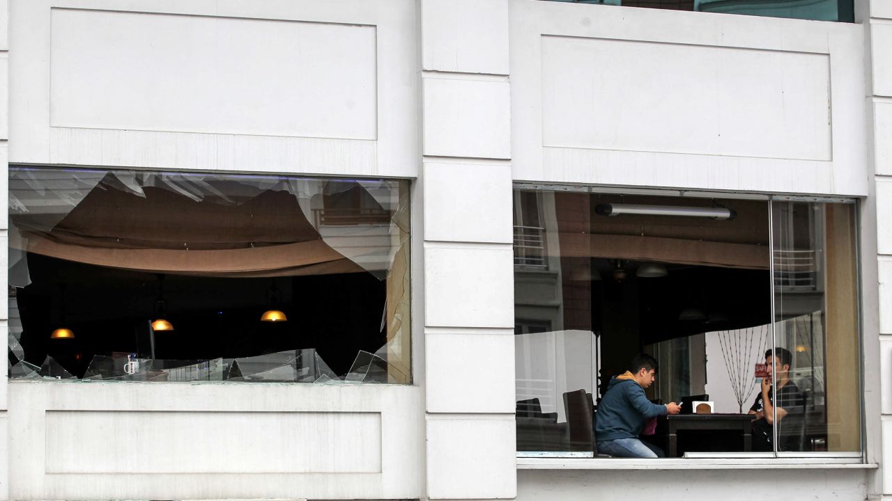 Two men sit near the broken windows of a restaurant as police officers secure the area.
