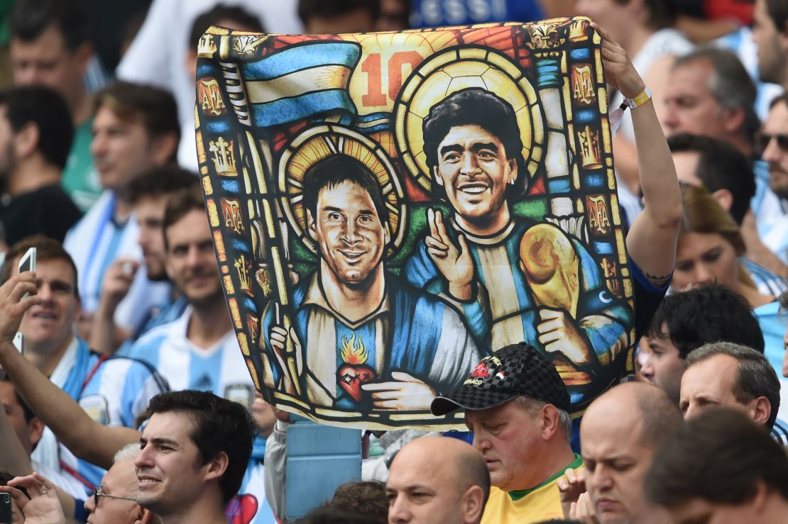 Lionel Messi and Diego Maradona depicted as saints on a supporter's flag. Maradona holds the World Cup trophy while Messi, crucially, does not. 