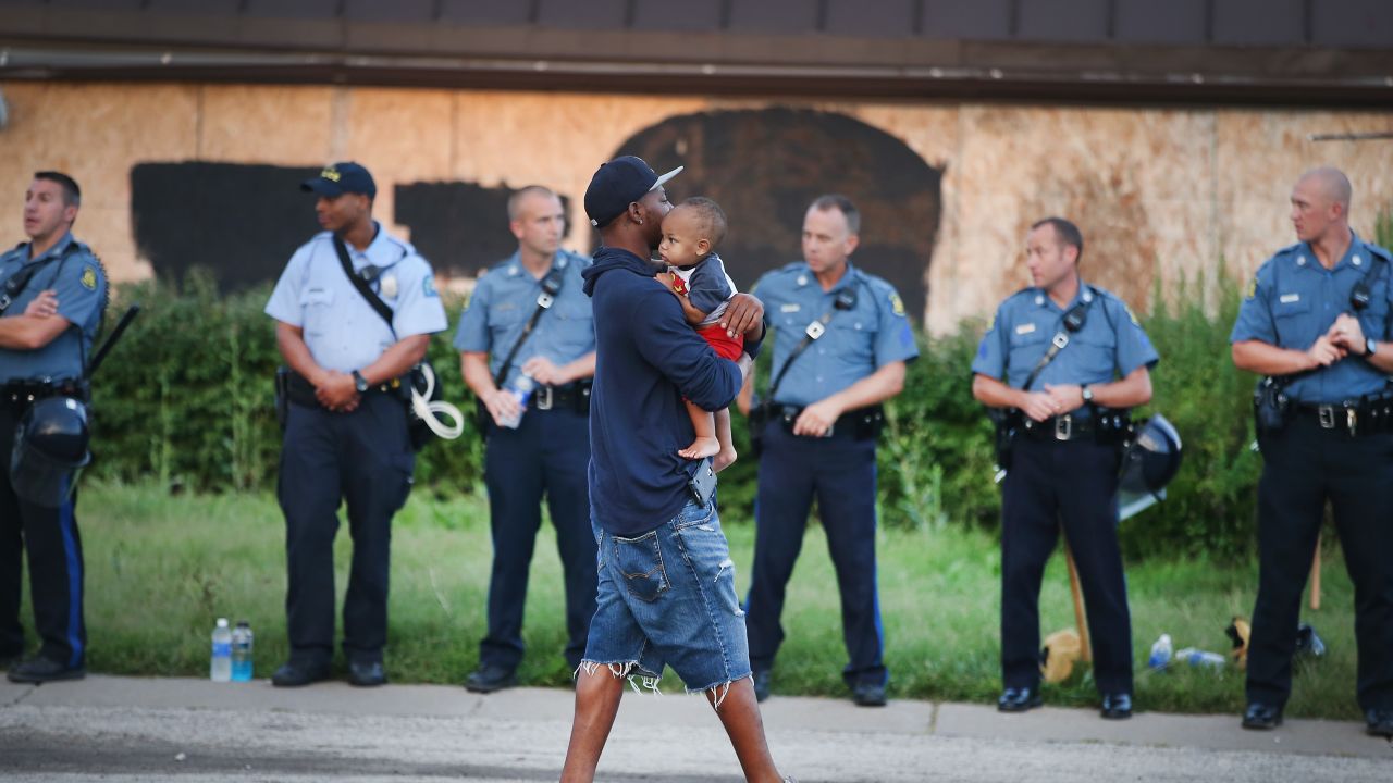 FERGUSON, MO - AUGUST 17: A man carries his child past a row of police tasked with keeping the peace as demonstration to protest the killing of teenager Michael Brown on August 17, 2014 in Ferguson, Missouri.   Despite the Brown family's continued call for peaceful demonstrations, violent protests have erupted nearly every night in Ferguson since his death. (Photo by Scott Olson/Getty Images)