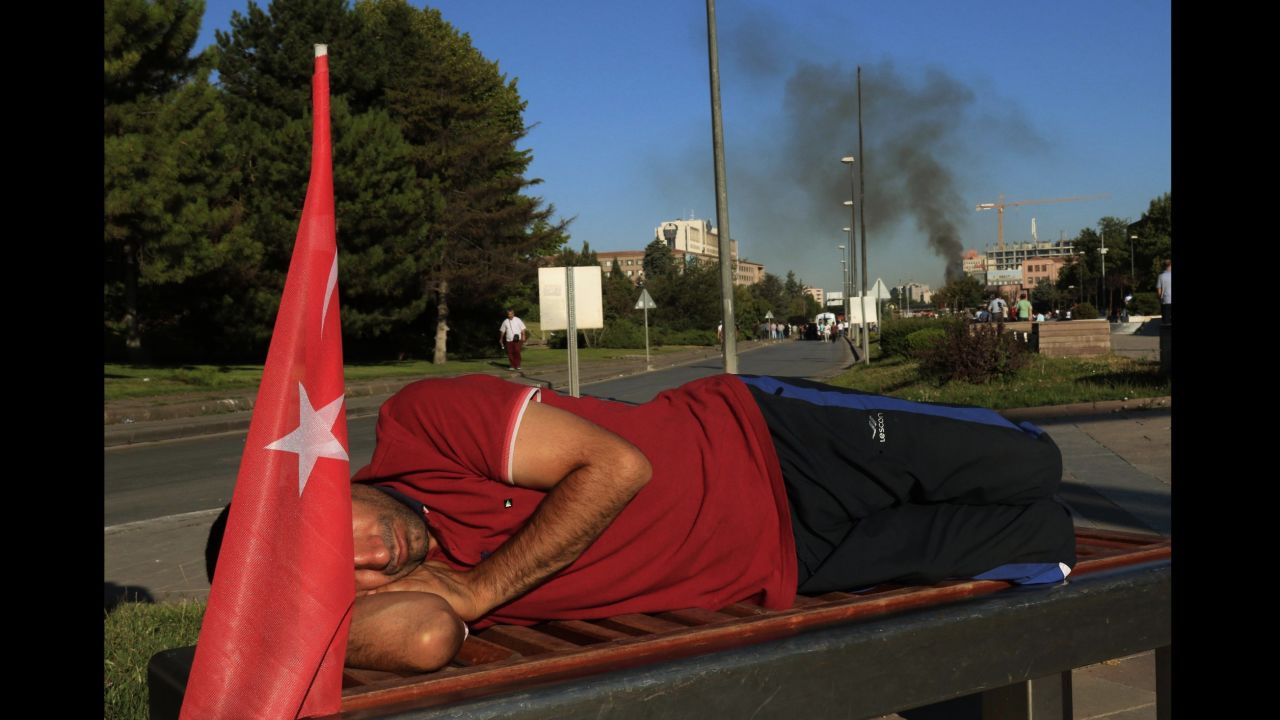 A protester rests on a bench as smoke billows from the Turkish military headquarters in Ankara.  
