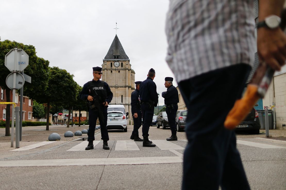 A man carries a French baguette as police officers stand guard the Saint-Etienne church.