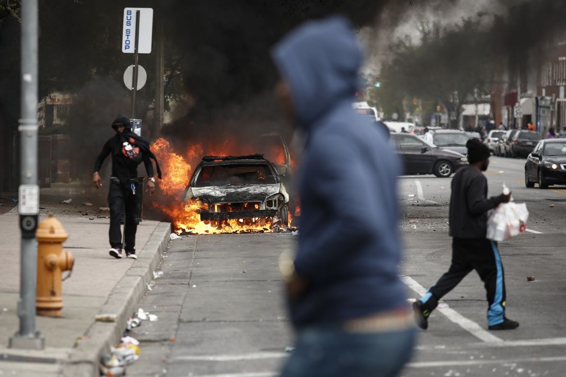 People walk past burning cars near the intersection of Pennsylvania Avenue and North Avenue in Baltimore on April 27, 2015. Riots erupted in the city following the funeral service for Freddie Gray, who died while in Baltimore Police custody.