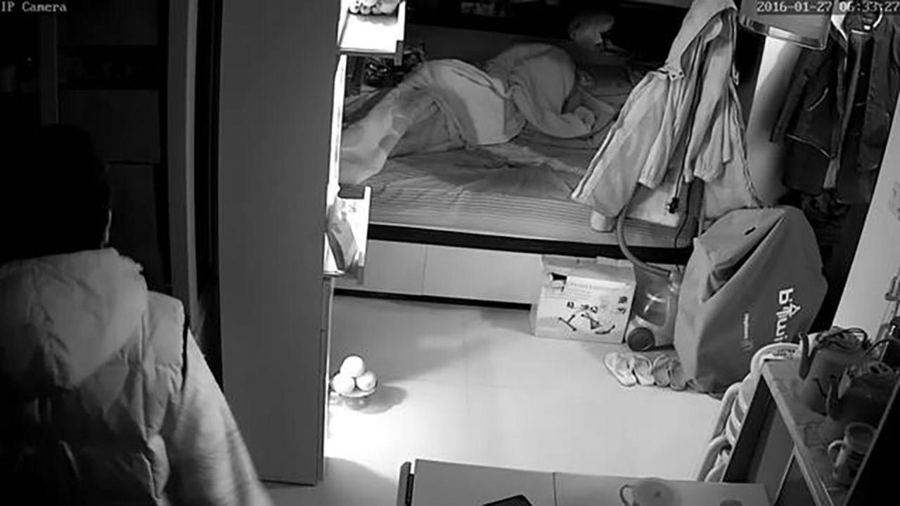 A figure stands to the left of the photo near to a bed with crumpled covers. 