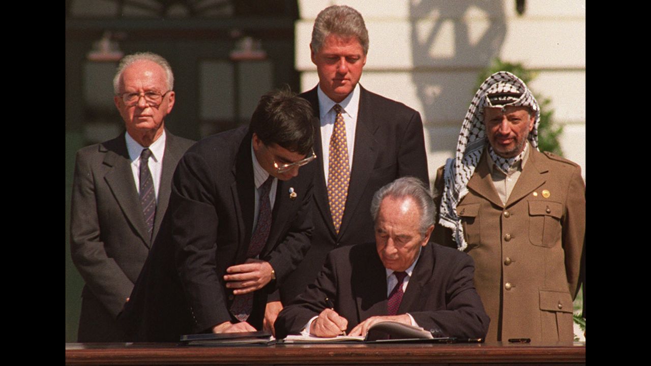 Israeli Foreign Minister Shimon Peres, with an unidentified aide, signs a peace agreement on September 13, 1993 between Israel and the Palestine Liberation Organization (PLO) in Oslo, Norway. Israeli Prime Minister Yitzhak Rabin, President Bill Clinton and PLO Chairman Yasser Arafat look on from behind. 