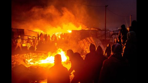 Migrants stand next to a burning shack in the camp on Tuesday, March 1.