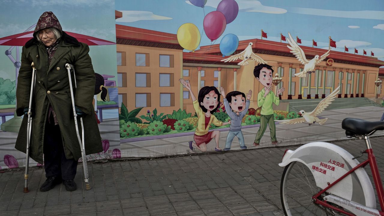 BEIJING, CHINA - DECEMBER 1: A disabled Chinese pensioner stands in the street next to a propaganda billboard showing a cartoon scene depicting the Great Hall of the People, in the street on December 1, 2014 in Beijing, China. Though China's economy is one of the fastest growing in the world and the standard of living has dramatically risen for millions in recent years, there are still many urban poor in the cities who are often migrants from rural areas. (Photo by Kevin Frayer/Getty Images)