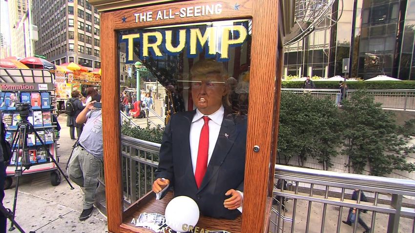Meet Trump the Fortune Teller. Jeanne Moos reports animatronic Trump is attracting crowds.    Fortune Teller Trump   The Donald now has a crystal ball and is telling fortunes. The All Seeing Trump is popping up in front of places like Trump Tower and Fox News. And on Wednesday we got a chance to fool around with him as he attracted a crowd of laughing spectators outside the Trump International Hotel. He's an animatronic likeness of the Donald inside a booth. He makes funny predictions about the future (the predictions mock the Donald's positions) and hands out written "misfortunes." He's modeled after "Zoltar", the fortune teller in the Tom Hanks' movie "Big." The All Seeing Trump was the brain child of anonymous Brooklyn artists and is voiced by a comedian impersonating Trump.  Cost to build it...$9000. When they set it up outside Trump Tower, there's video of clueless security looking at it in bewilderment. Finally the police turned off the sound to shut it up. Click link below to hear it.