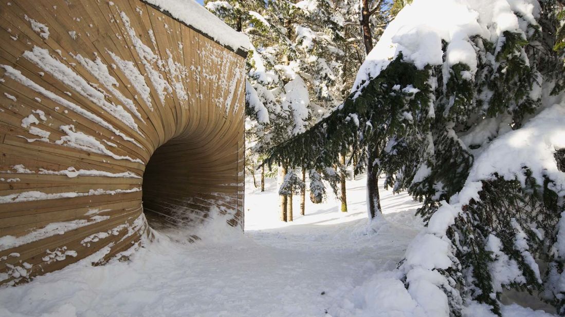 This serene residence features a curved tunnel at its entrance, which is made from bent planks of raw larch wood. The tunnel appears like a wooden vortex hidden behind trees. 