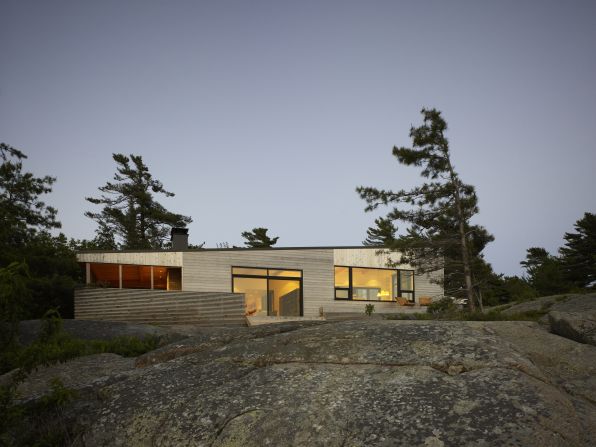 Superkül architects' Shift Cottage sits on the shore of Georgian Bay in Ontario, Canada. The cottage's windows overlook a row of conifer trees, and a cedar deck offers ample outdoor living space. 