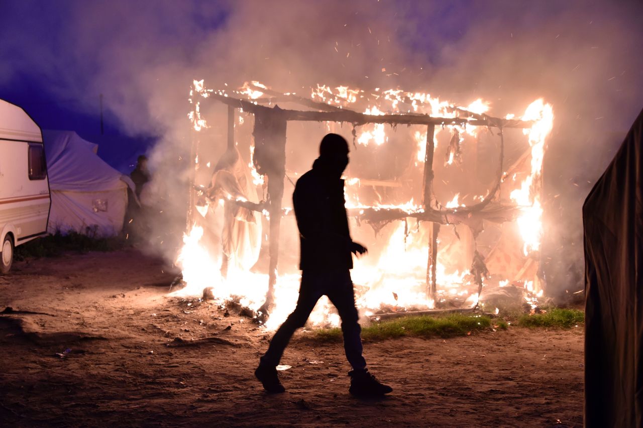 A migrant walks past a burning shack that was set on fire, as a demolition crew began tearing structures down on Tuesday, October 25.
