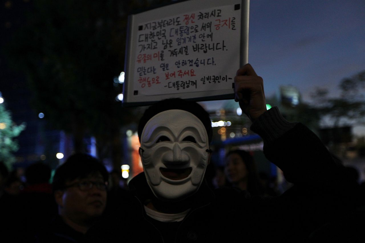 A protester wearing a traditional Korean mask holds a placard saying "You Need to Resign Now" during the protest. 