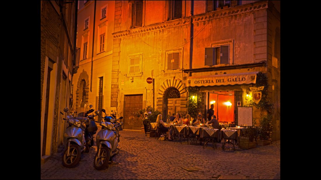 A typical night time corner of the city with an outdoor cafe, diners and parked scooters.  