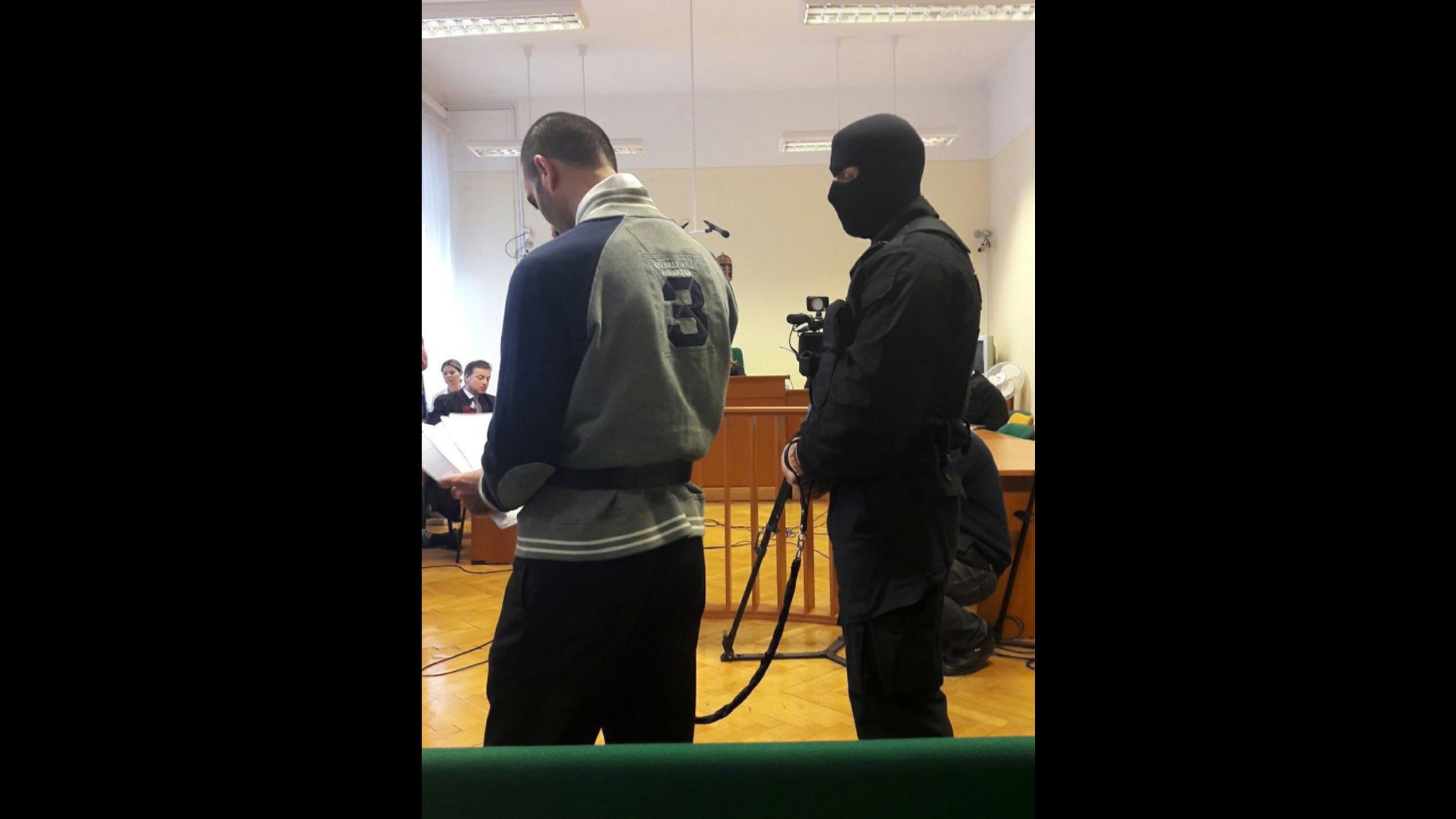 Ahmed H. stands in court on November 30, 2016, flanked by a balaclava-wearing guard. 