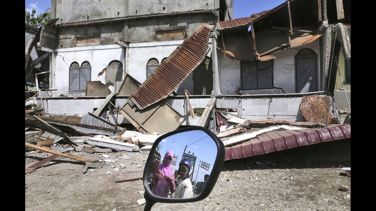 Two women are reflected in the mirror of a motorbike parked near a damaged building in Pidie Jaya.