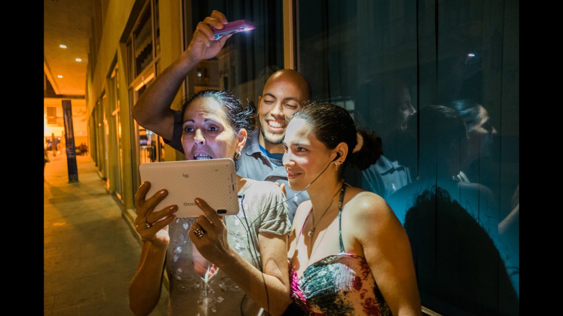 Leonor Cuza Tamayo, 52, chats with a sister in Miami as her son, Jose Luis Rodriguez, 33, holds a light over her head. 