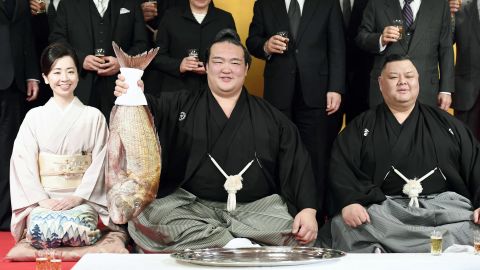 Kisenosato holds a fish during a ceremony promoting him to the highest rank of sumo wrestling, accompanied by his trainer and trainer's wife. 