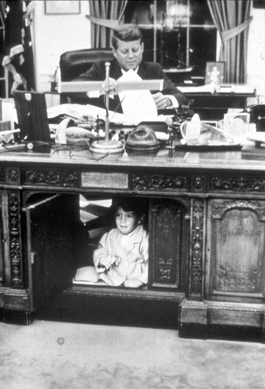John F. Kennedy works at the Resolute Desk in the Oval Office while his son, John Jr., peeks out from the panel underneath the desk.