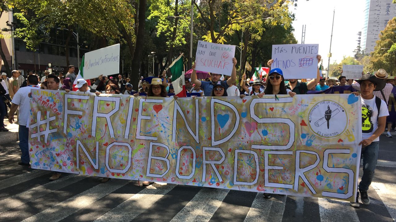 Protesters hold a banner that reads "#Friends, No Borders." Additional signs from left to right: "#friendship with no borders," "to govern you have to listen," "Less walls."
