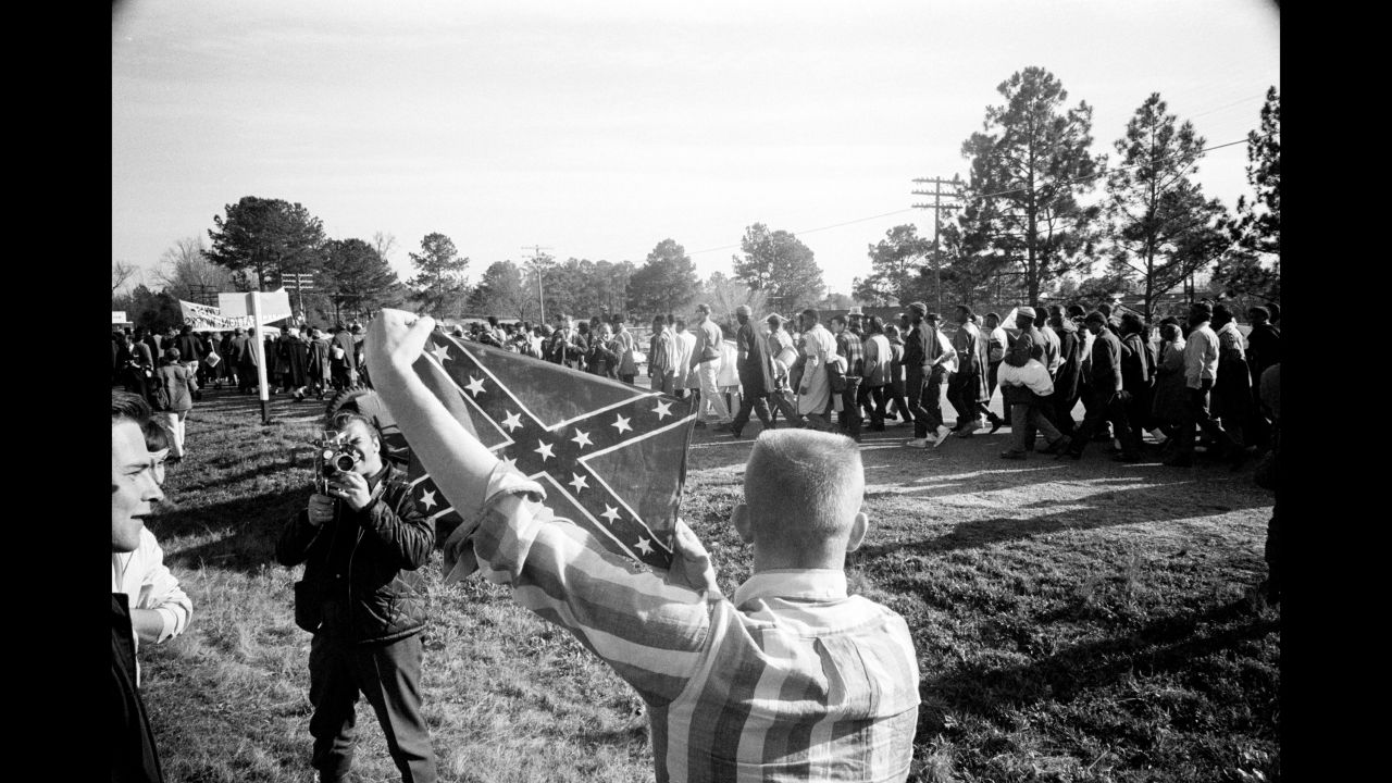 Marchers walk past a young white man holding a Confederate battle flag.