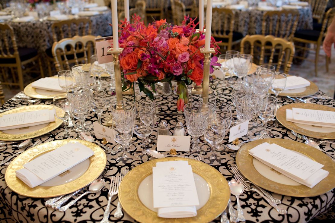White House State Dining Room 2010. Black and white-patterned linens and bright coral flowers create an elegant backdrop with table décor selected from the historic White House collection: gold-rimmed Clinton base plates, coral-red Reagan china, Charleston candlesticks, King Charles flatware, and eagle motif place card holders. Illuminated with votive and taper candles and mirrored centerpiece containers capturing the candlelight and flowers.