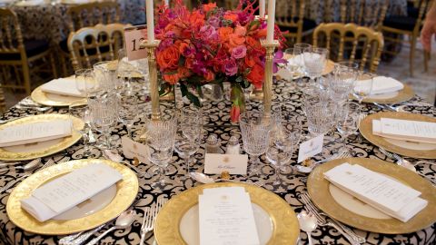 White House State Dining Room 2010. Black and white-patterned linens and bright coral flowers create an elegant backdrop with table décor selected from the historic White House collection: gold-rimmed Clinton base plates, coral-red Reagan china, Charleston candlesticks, King Charles flatware, and eagle motif place card holders. Illuminated with votive and taper candles and mirrored centerpiece containers capturing the candlelight and flowers.