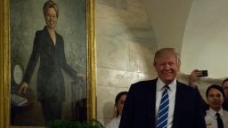 President Donald Trump walks in a corridor of the White House to greet visitors while a portrait of Hillary Clinton hangs on the wall on March 7, 2017.