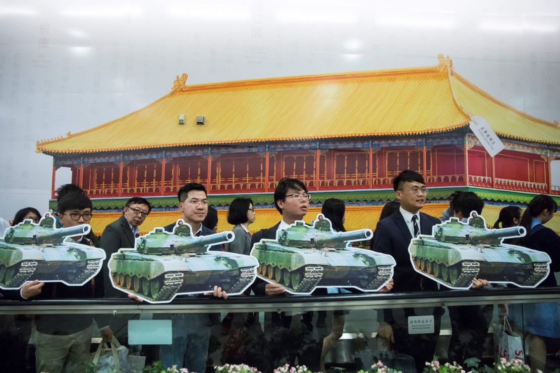 Protesters hold pictures of tanks in front of a giant advert for the Hong Kong Palace Museum.