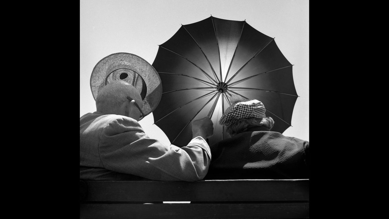 A man holds up an umbrella and a hat while sitting on a Coney Island bench in 1948.