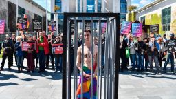 An activist stands naked, wrapped in a rainbow flag, in a mock cage in front of the Chancellery in Berlin on April 30, 2017, during a demonstration calling on Russian President to put an end to the persecution of gay men in Chechnya. 
The protestors called on German Chancellor Angela Merkel, who will meet Putin in Sochi on May 2, 2017, to raise the issue with him. / AFP PHOTO / John MACDOUGALL        (Photo credit should read JOHN MACDOUGALL/AFP/Getty Images)