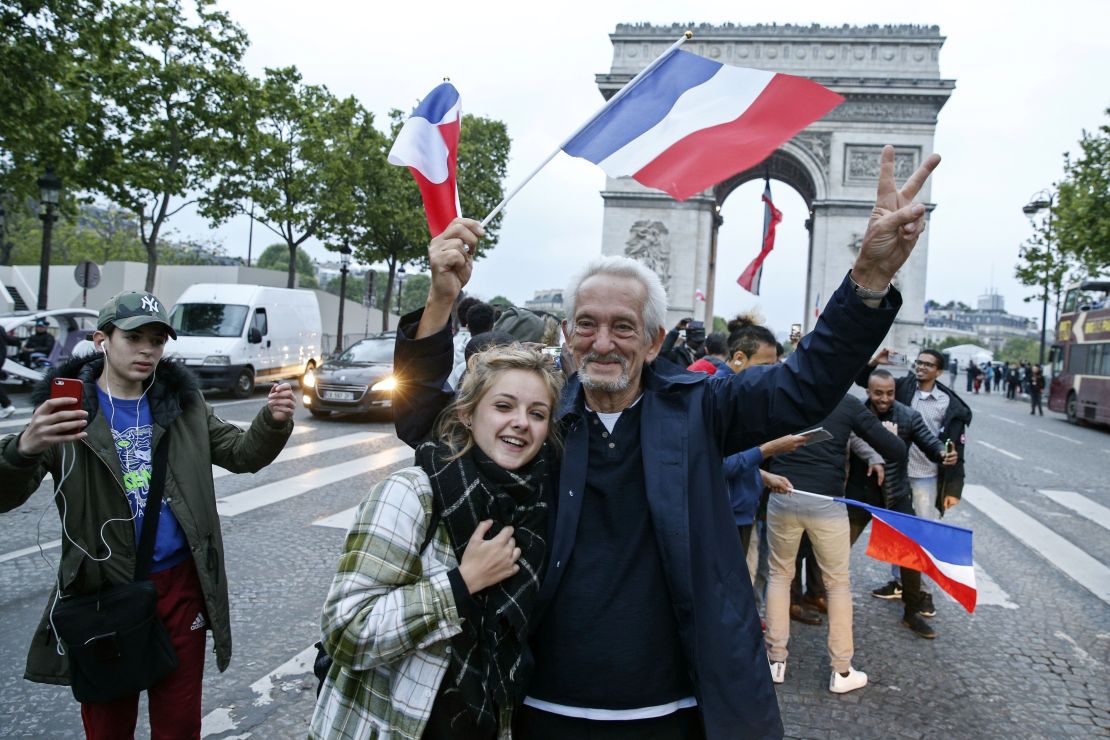 A man, flanked by a young woman, waves a French national flag next to people gesturing and shouting on the French avenue of the Champs Elysees by the Arc de Triomphe.