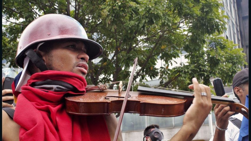 A young protester plays his violin in the streets of Caracas as national guards throw teargas and pellets.