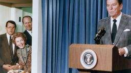 4th February 1983:  US President Ronald Reagan stands behind a lectern as First Lady Nancy Reagan stands in a side doorway, planning to surprise the President with a birthday cake, at the White House, Washington, DC. Two men stand behind the First Lady.  