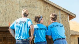 A group of three volunteers helping to build a house for a family in need.  One woman stands in the middle between two men.   Their backs are to the camera, showing the word VOLUNTEER written on the back of their blue shirts.