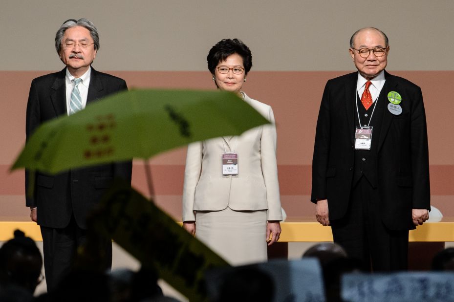 Carrie Lam (center) stands on stage after being selected to be the next Hong Kong Chief Executive on March 26, 2017. In the foreground, a yellow umbrella is unfurled to protest the undemocratic nature of the race Lam won. 