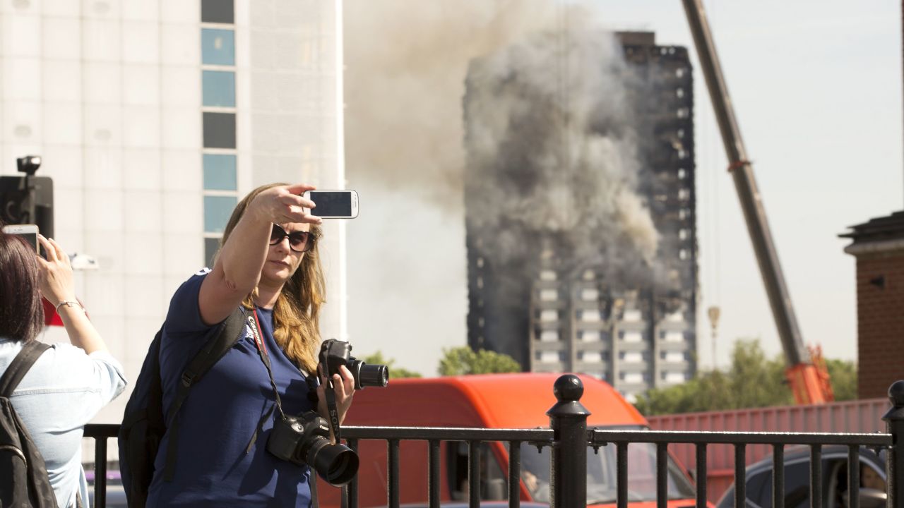 A woman takes a selfie as pedestrians stop to watch flames being put out at Grenfell Tower in the distance
