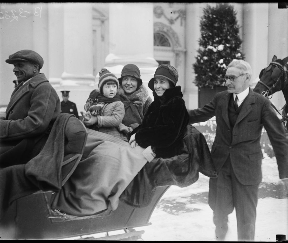American First Lady Grace Coolidge (nee Goodhue, 1879 - 1957) (center, in dark coat) smiles with unidentified others as she sits in a sleigh, while Chief White House Usher Ike Hoover (1871 - 1933) (right), walks beside them outside the White House, Washington DC, 1929. 