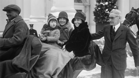 American First Lady Grace Coolidge (nee Goodhue, 1879 - 1957) (center, in dark coat) smiles with unidentified others as she sits in a sleigh, while Chief White House Usher Ike Hoover (1871 - 1933) (right), walks beside them outside the White House, Washington DC, 1929. 