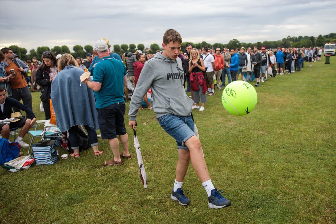 A young man performs kick-ups with a giant tennis ball as tennis fans wait in line. 