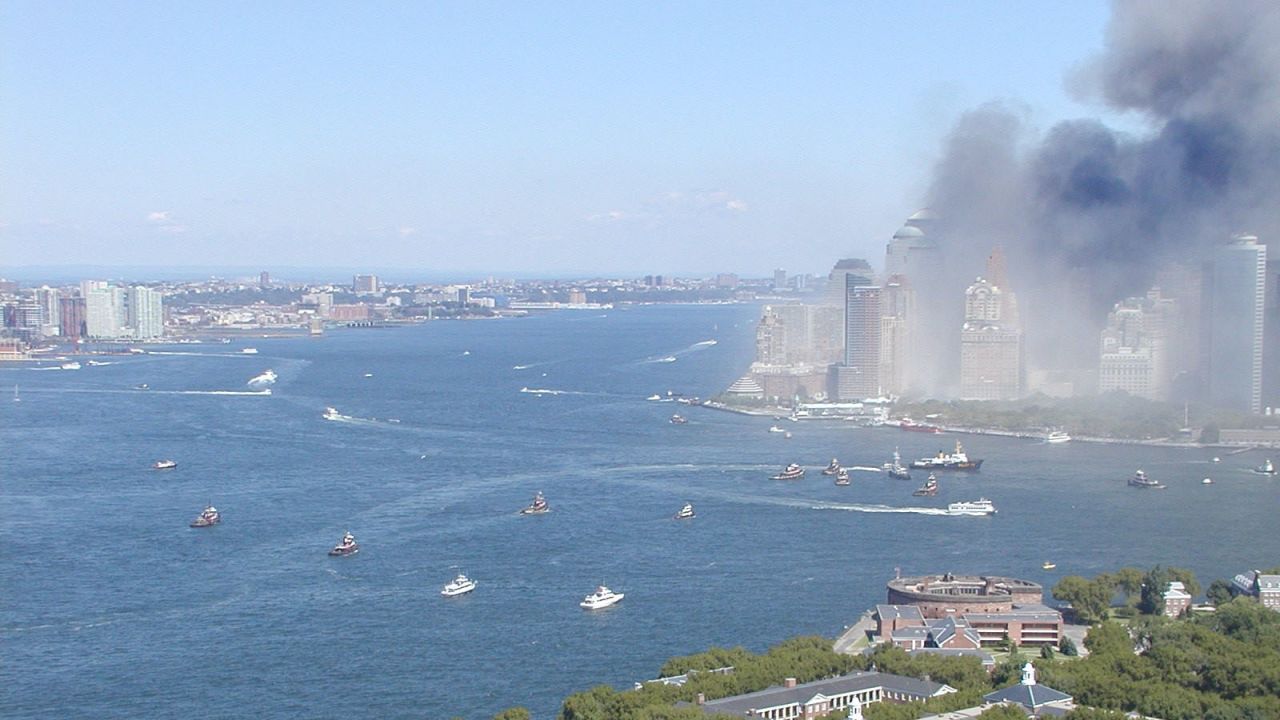 The view north from above Governor's Island shows a fleet of diverse vessels approaching smoke-filled Lower Manhattan and the Battery at the southern tip of the island. New Jersey is on the left.