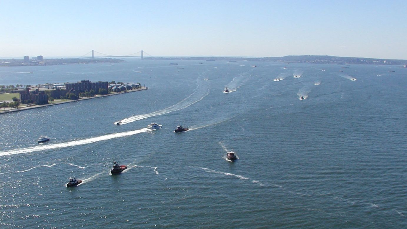 The view south from Manhattan shows tugs racing from Staten Island and New Jersey to the Battery. On the left is the southern tip of Governors Island. The Verrazano-Narrows Bridge is visible in the background. 