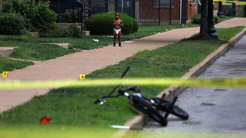 A baby stands outside a home in St. Louis as police investigate the scene of a fatal shooting on Monday, August 7.