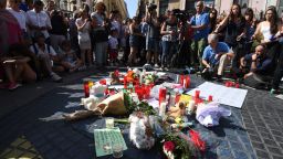 People stand next to flowers, candles and other items set up on the Las Ramblas boulevard in Barcelona as they pay tribute to the victims of the Barcelona attack, a day after a van ploughed into the crowd, killing 13 persons and injuring over 100 on August 18, 2017. 